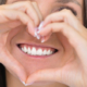 Frau zeigt beim Lächeln die Zähne und formt mit ihren Händen ein Herz vor dem Mund