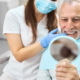 Älterer Mann auf einem Zahnarztstuhl schaut mit offenem Mund in einen Handspiegel. Hinter ihm sitzt eine frau, die mit der einen Hand auf seine Zähne zeigt.